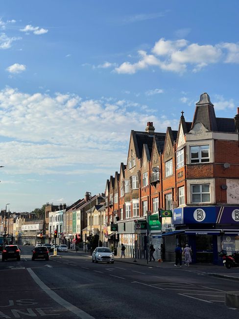 The image shows a typical street scene in Catford with a row of multi-storey brick and plaster residential buildings and shops lining the pavement. Several cars are parked along the road while others 