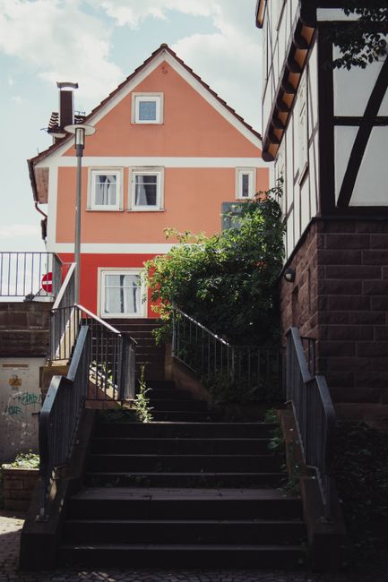 A narrow outdoor stairway with stone steps ascending between two brick buildings in an urban setting. The left side of the alley features a black metal handrail and a wall-mounted lantern, while the r